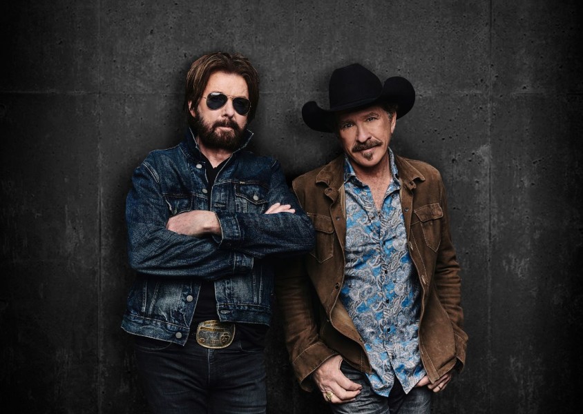 Brooks & Dunn stood against a black wall one wearing a denim jacket and black sunglasses, the other a brown jacket and black cowboy hatx